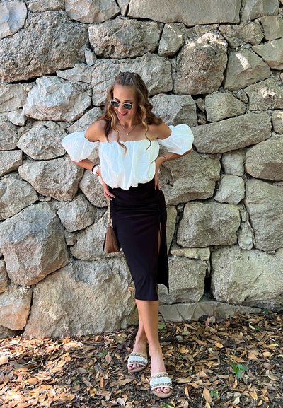 Blusa blanca fuera de los hombros con mangas voluminosas, combinada con una falda midi negra con una abertura lateral. Llevada con sandalias beige adornadas.