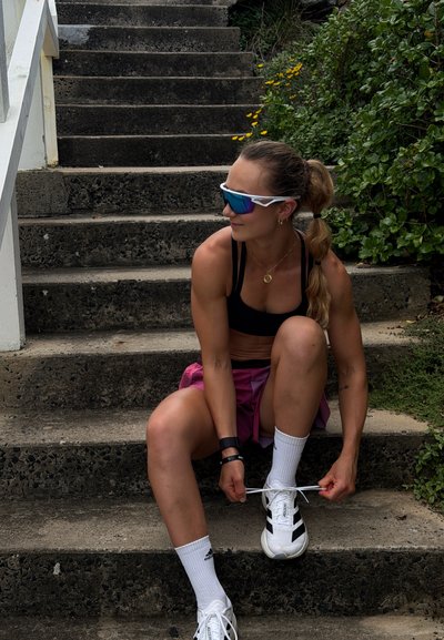 Mujer atlética con gafas de sol deportivas sentada en escalones al aire libre atándose unas zapatillas blancas, rodeada de vegetación y flores amarillas.