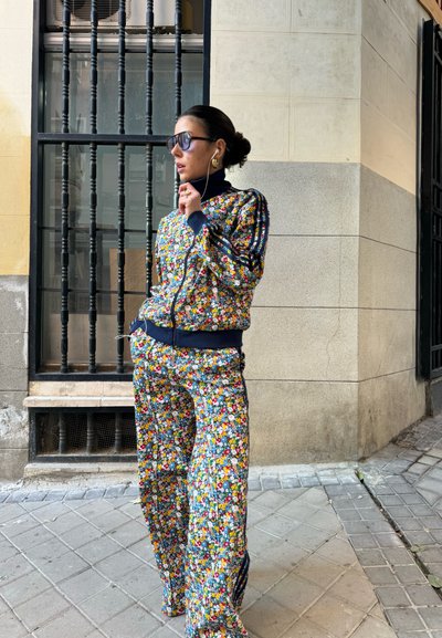 Chándal de estampado floral en azul, verde, rojo y amarillo, con cierre de cremallera en el frente, cuello alto y rayas deportivas en los laterales. Lleva gafas de sol.