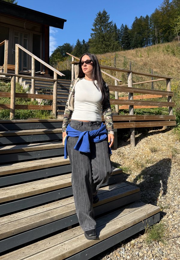 Mujer con gafas de sol y mangas de camuflaje baja por escaleras de madera al aire libre con una cabaña y árboles al fondo en un día soleado.