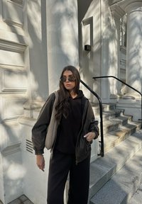 Καφέ δερμάτινη μπομπέρ ζακέτα πάνω από μια μαύρη μπλούζα φούτερ, σε συνδυασμό με μαύρα παντελόνια φαρδιάς γραμμής και γυαλιά ηλίου στρογγυλής μορφής. Η πλεκτή τσάντα ώμου προσθέτει υφή.