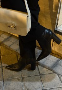 Stivali ankle in pelle nera con punta affusolata, tacchi a blocco e texture liscia, abbinati a una borsa color crema con chiusura in metallo.