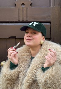 Persona con cappotto beige peloso tiene un rossetto argento e mostra un orecchino dorato con texture. Indossa un berretto di velluto a coste verde con "NY" bianco.