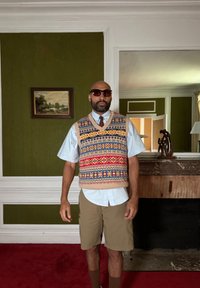 Πλεκτό γιλέκο σε μπεζ χρώμα με πολύχρωμα γεωμετρικά σχέδια, φορεμένο πάνω από ένα ανοιχτό μπλε πουκάμισο. Συνδυασμένο με καφέ σορτσάκι και γυαλιά ηλίου, στέκεται σε εσωτερικό χώρο.