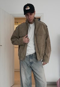 Jeune homme à l'intérieur portant une casquette noire avec un patch d'ange, une veste en daim beige par-dessus une chemise blanche, et un jean délavé et ample, debout près d'une porte.