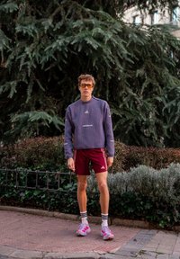 Jonge man draagt een paarse trui, wijnrode korte broek, witte sneakers en oranje zonnebril, staand op de stoep bij groenblijvende planten.