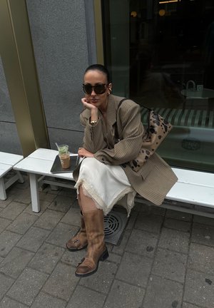 Braun karierter Blazer über einem cremefarbenen Rock mit Spitzenbesatz, kombiniert mit hohen braunen Lederstiefeln. Eine gemusterte Handtasche und ein Eiskaffee auf einem Tablett.