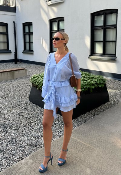 Mujer con vestido azul claro con volantes y sandalias de tacón alto, llevando un bolso marrón al hombro, de pie en un patio de piedra cerca de un edificio blanco.