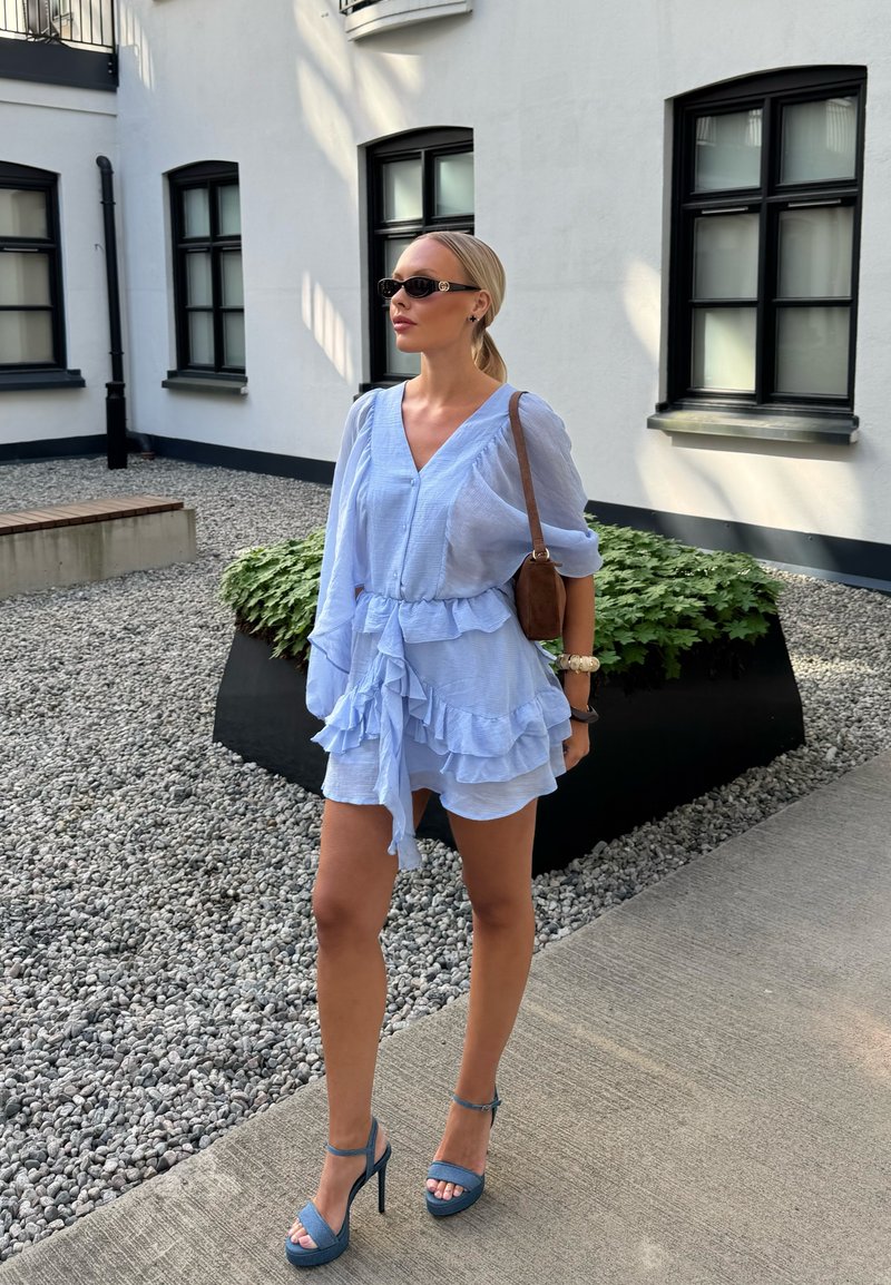 Femme portant une robe bleue claire à volants et des sandales à talons hauts, portant un sac à épaule marron, debout dans une cour en pierre près d'un bâtiment blanc.