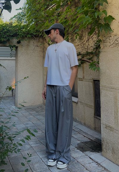 Camisa blanca de manga corta, pantalones gris de pierna ancha y zapatillas en blanco y negro. El escenario presenta un pavimento de piedra y una pared texturizada con vegetación.