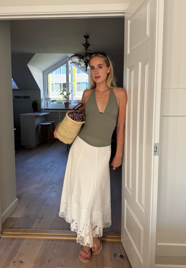 Donna con top oliva senza maniche e gonna bianca che tiene un cestino intrecciato, in piedi sull'uscio di una stanza luminosa con pavimento in legno.