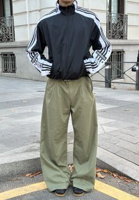 Black zip-up jacket featuring white stripes and a logo; loose-fitting green trousers with pleats; black shoes with tan soles, standing outdoors.