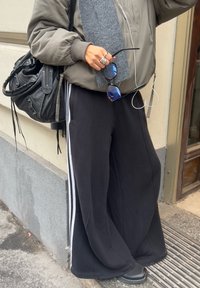 Černé oversized tepláky s bílými bočními pruhy, spárované se šedou bomber bundou, držící šedou látku a sluneční brýle, s černou taškou.