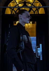 Musta paita, jossa on helmikoristeinen reunus, yhdistettynä pieneen mustaan käsilaukkuun. Taustalla on tummansävyinen ovi ja chandelier.