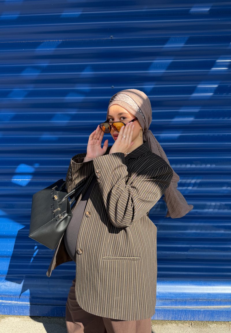 Femme enceinte portant un foulard et un blazer oversize à fines rayures ajuste ses lunettes de soleil, tenant un sac à main noir devant un fond métallique ondulé bleu.