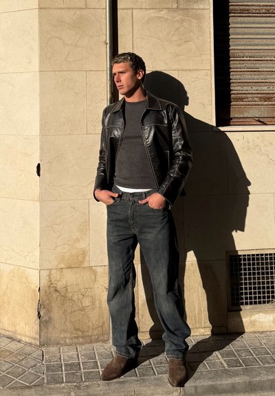 Hombre con chaqueta de cuero negra, jeans oscuros y botas marrones apoyado contra una pared beige iluminada por el sol con las manos en los bolsillos.