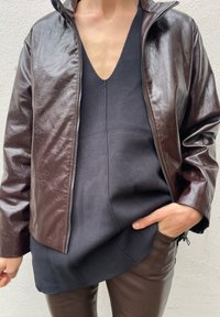 Brązowa skórzana kurtka zapinana na zamek błyskawiczny z wysokim kołnierzem, noszona na czarnej bluzce. Kurtka ma gładką fakturę i lekko błyszczące wykończenie.
