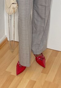 Sapatos de couro verniz vermelho com bicos finos e fivelas prateadas; combinados com calças cinza claro em padrão xadrez. O fundo apresenta um chão de madeira.