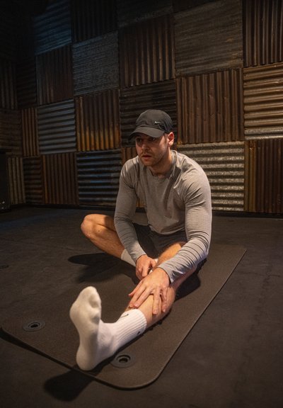 Camisa atlética de manga larga gris y pantalones cortos negros llevados por una persona que se estira sobre una esterilla de ejercicio negra texturizada en un entorno de gimnasio rústico.