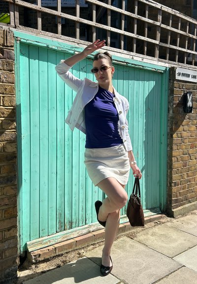 Mujer posando con gafas de sol, chaqueta blanca, top azul marino y falda beige, de pie sobre una pierna frente a una puerta de madera turquesa bajo la luz del sol.