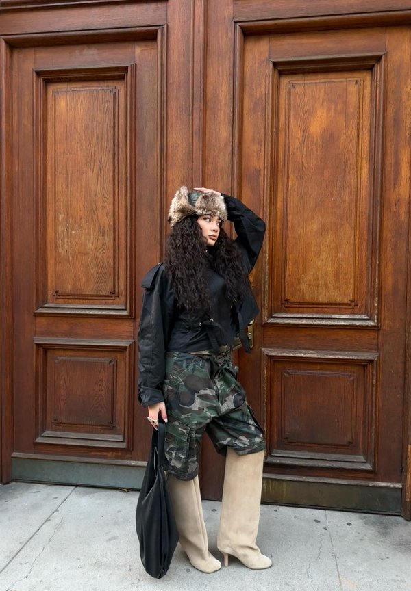 Giacca nera corta, pantaloni cargo mimetici, stivaletti beige con tacco a stiletto e cappello foderato in pelliccia. In posa vicino a una porta in legno con una borsa nera.