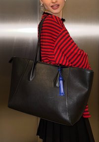 Personne portant une chemise rayée rouge et noire, portant un grand sac fourre-tout en cuir noir avec un porte-clés bleu, sur un fond métallique.