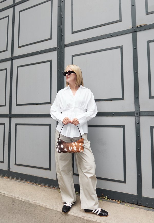 Žena s blond vlasy, která má na sobě sluneční brýle, bílou košili, béžové kalhoty, černo-bílé boty a drží hnědou tašku s tečkami, stojí u šedé panelové stěny.