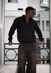 Sweatshirt zippé noir avec col côtelé et manches longues, associé à un pantalon sombre ample. Le modèle s'appuie sur un garde-corps de balcon.