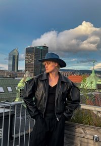 Μαύρο δερμάτινο μπόμбер τζάκετ με φουσκωμένα μανίκια, φορεμένο πάνω από μια μαύρη τοπ. Μπλε καπέλο με φαρδύ γείσο, αστικό φόντο με κτίρια και πράσινο.