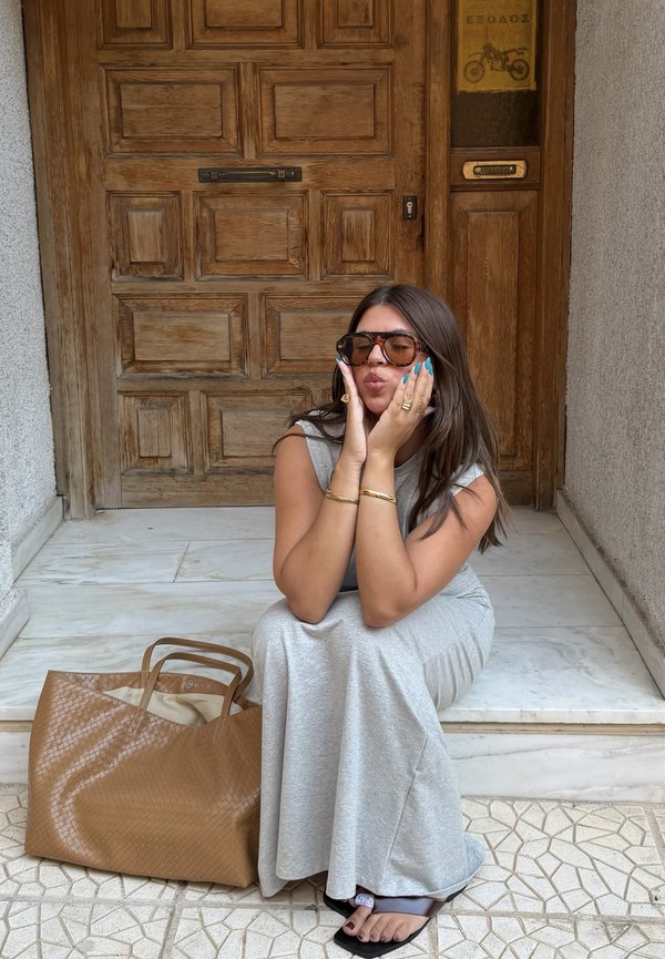 Donna con lunghi capelli castani e occhiali da sole seduta sui gradini davanti a una porta di legno, indossa un vestito grigio e sandali neri, con una grande borsa marrone nelle vicinanze.