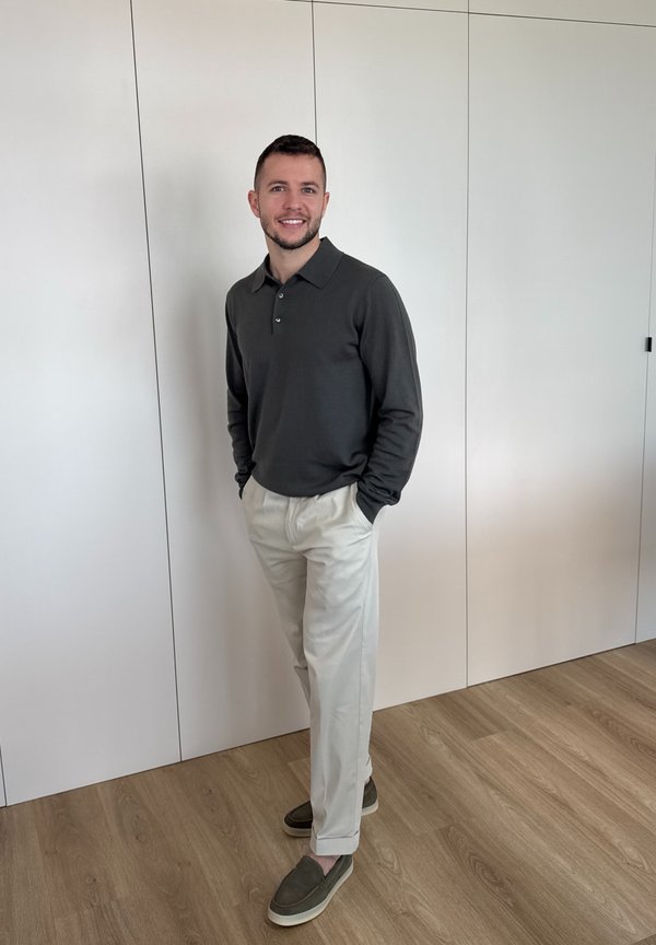 Mand stående indendørs med hænderne i lommerne, iført en mørk langærmet polo, lyse bukser og mørke slip-on sko.
