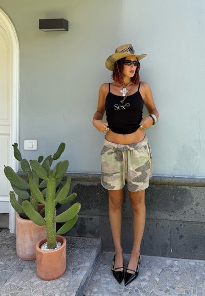 Top negro sin mangas con el texto "Sex©", pantalones cortos de camuflaje y zapatos negros de punta. Un sombrero de paja y gafas de sol completan el look al aire libre. Un cactus está cerca.