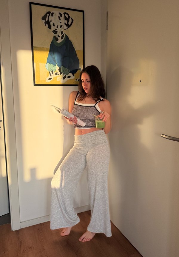 Młoda kobieta w casualowych ubraniach czytająca magazyn i trzymająca zielone smoothie, stojąca boso obok ściany z obrazem psa.