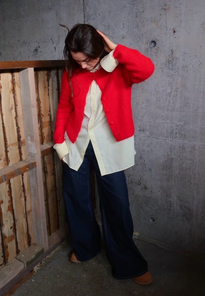 Cárdigan de punto rojo sobre una camisa de botón crema, combinado con jeans de mezclilla oscuros de pierna ancha y calzado marrón. Evidentes materiales texturizados.