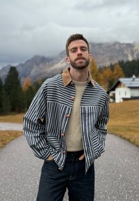 Randig jacka med brun krage, knappstängning och sidofickor. Buren över en ljus tröja och mörka jeans mot en bergig bakgrund.