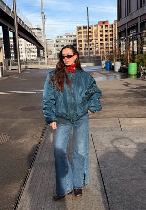Mujer joven con chaqueta bomber azul extragrande, bufanda roja, vaqueros de pierna ancha y gafas de sol, de pie en una calle de la ciudad con edificios y barriles detrás.