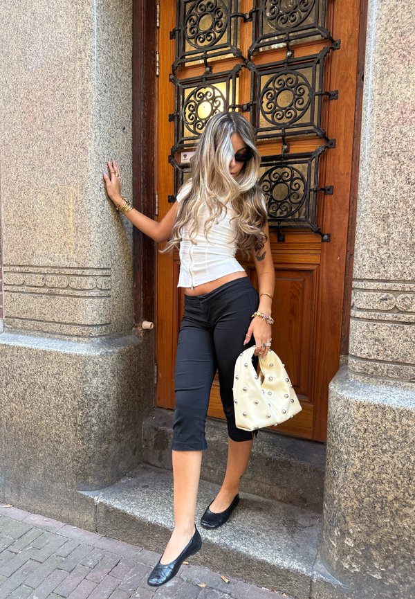 Femme aux longs cheveux blonds portant des lunettes de soleil, un haut blanc, un pantalon capri noir, tenant un sac à main blanc orné de perles, debout près d'une porte en bois.