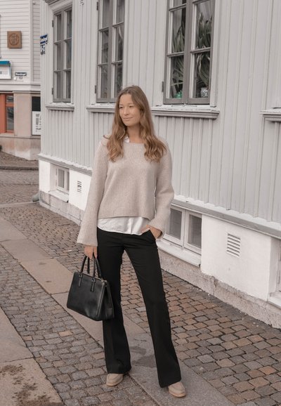 Suéter de punto beige claro sobre una camisa blanca, pantalones acampanados negros y calzado gris. Bolso negro. Calle de adoquines y edificios de madera al fondo.