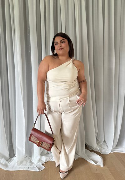 Top de un hombro color crema combinado con pantalones anchos plisados color crema. Sosteniendo un bolso marrón con un patrón de monograma. Textura suave.