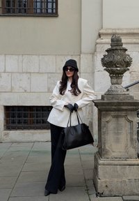 Blazer blanc, pantalon noir à coupe large, gants noirs et lunettes de soleil ; portant un sac à main noir. Fond en pierre grise avec une colonne ornée.