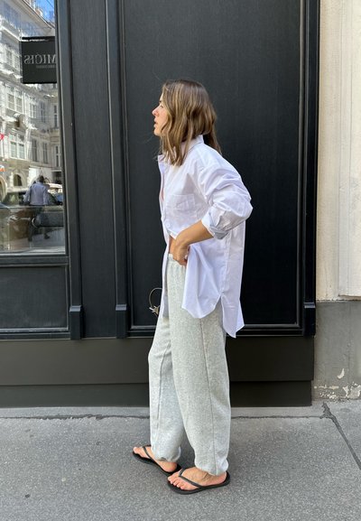 Camisa blanca de gran tamaño con botones, pantalones de chándal grises y chanclas negras. De pie junto a una pared negra, mirando hacia la izquierda, con una postura relajada.