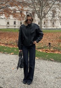 Črna usnjena bomber jakna s teksturirano površino, kombinirana z zvončastimi temnimi kavbojkami in okroglimi črnimi čevlji. Drži črno torbico.