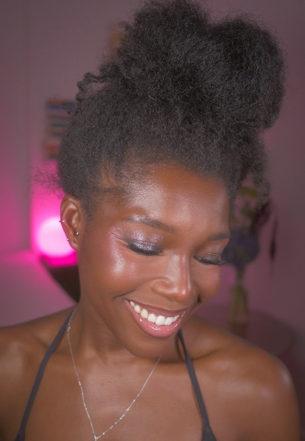 Une femme souriante aux cheveux naturellement bouclés, coiffés en hauteur, regarde vers le bas. Elle porte du maquillage, des boucles d’oreilles et un collier dans une pièce à l’éclairage doux.