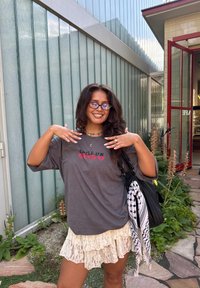 Tricou gri cu textul "SPILL UR GUTS" în roșu, fustă mini din dantelă stratificată, geantă neagră, ochelari și inele. Cadru exterior cu vegetație.