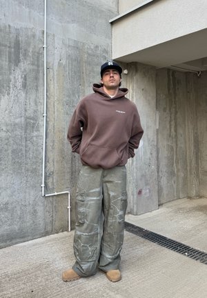 Sudadera marrón con un bolsillo frontal, pantalones cargo grises de corte holgado con textura y zapatos de lona color beige. Fondo de paredes de concreto.