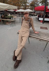 Beige overáll, alatta egy texturált, színes kardigán, barna fűzős csizmával, egy fa padon ülve egy szabadtéri piacon.
