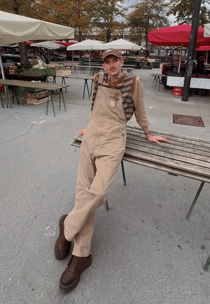 Overoles beige con un suéter texturizado y multicolor debajo, botas marrones con cordones, sentado en un banco de madera en un mercado al aire libre.