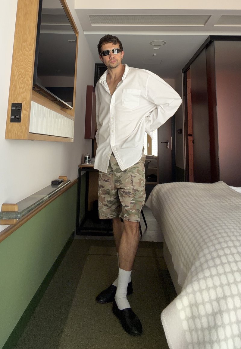 Homme portant des lunettes de soleil, une chemise blanche et un short camouflage se tient debout, la main derrière le dos, dans une chambre d'hôtel à côté d'un lit et d'une télévision.