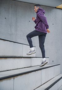 Träningsskor med grå och lila accenter, bärs med svarta avsmalnande byxor och en lila huvtröja, medan personen går uppför vita trappsteg.