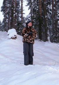 Een persoon staat in de sneeuw, gekleed in een bruin-zwart fleecejack met een patroon, zwarte cargobroek en handschoenen, met besneeuwde bomen op de achtergrond.